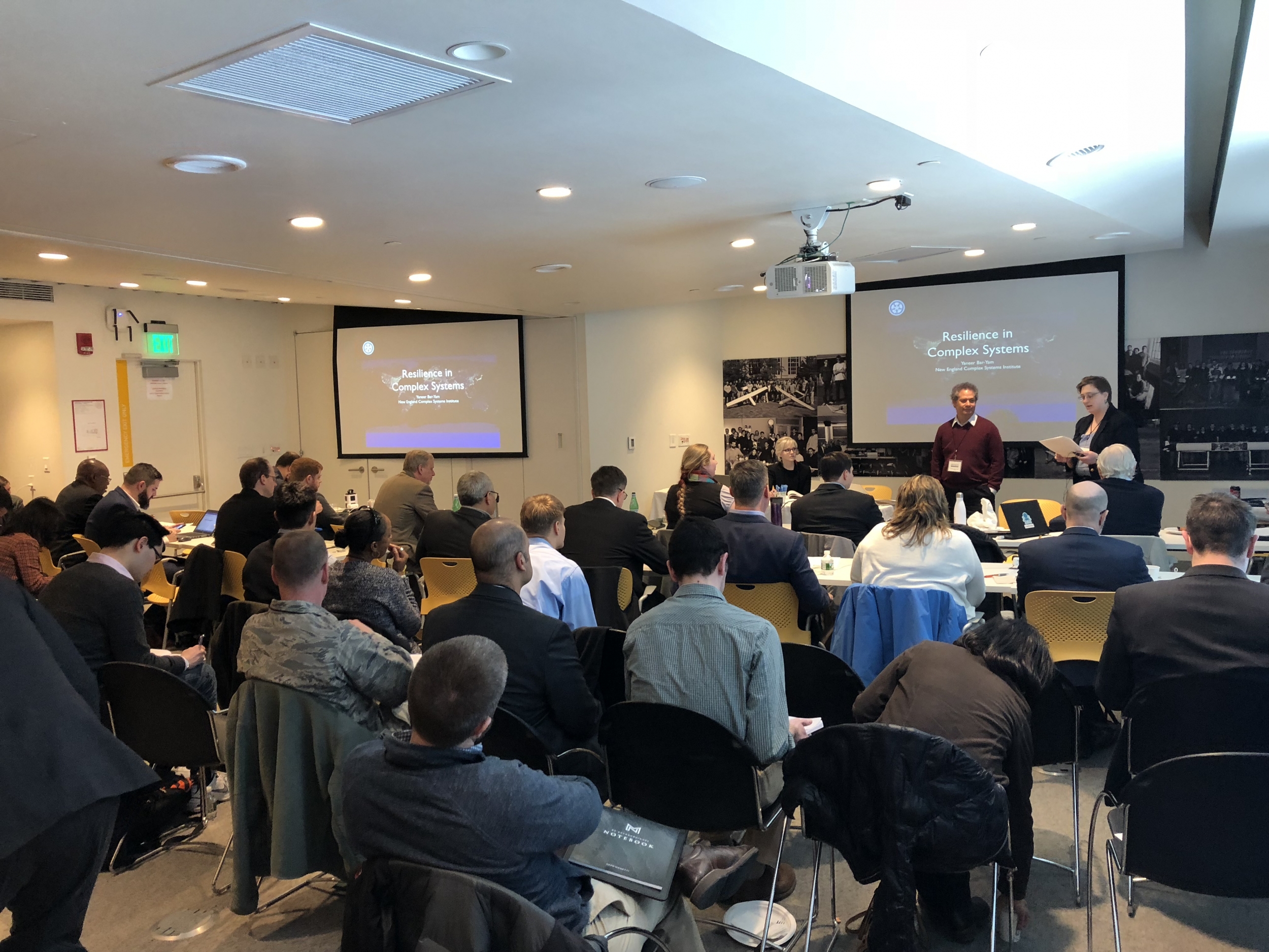 About thirty people sit in a classroom setting, with chairs facing a panel at front and projectors with a slide reading "Resilience in Complex Systems"