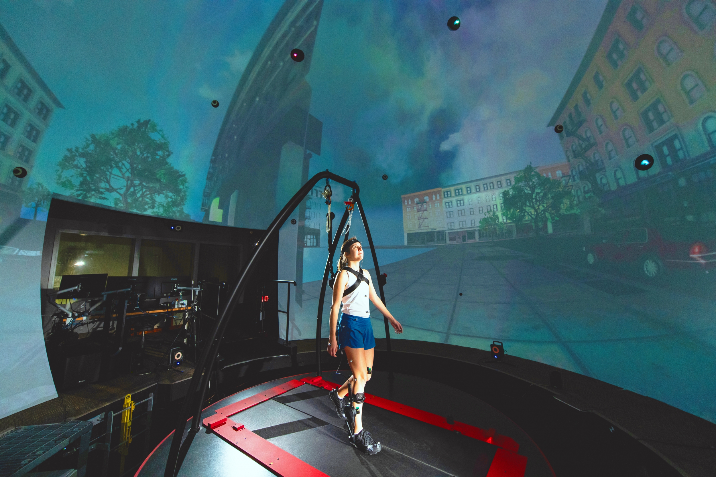 A woman, attached to a harness, stands on a treadmill, and is surrounded by a spherical screen displaying a city scene.