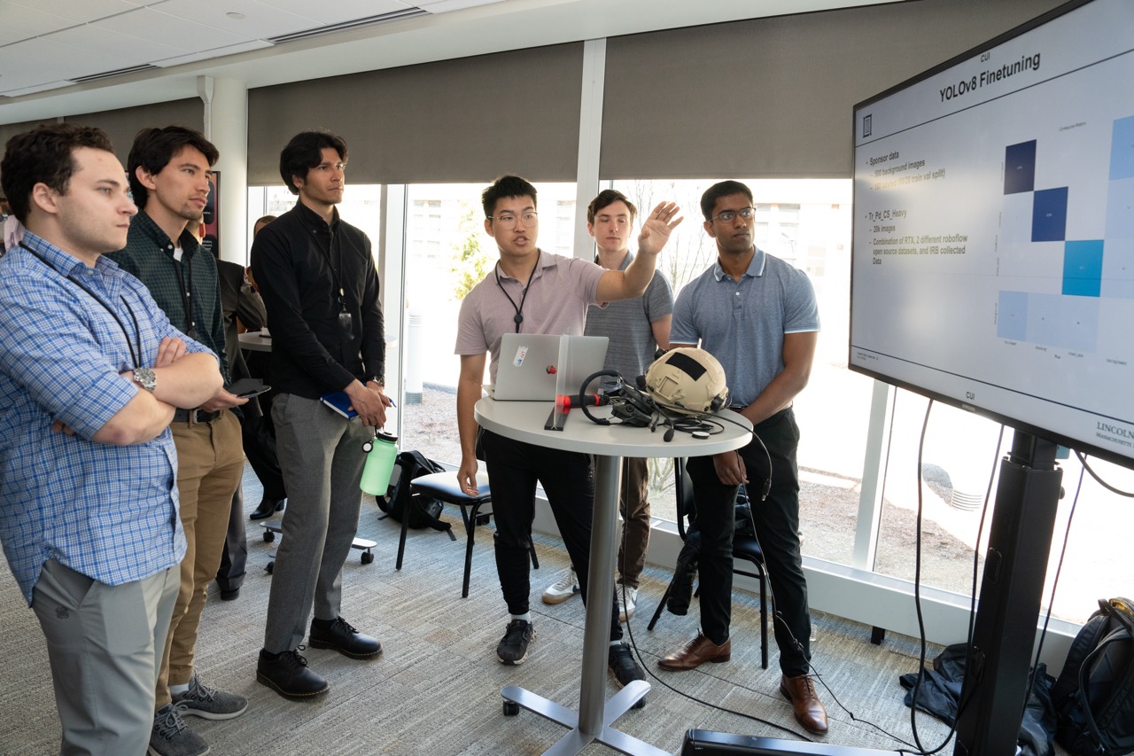 A group of cadets and staff look at a large monitor as a cadet presents his AI casualty status tool.