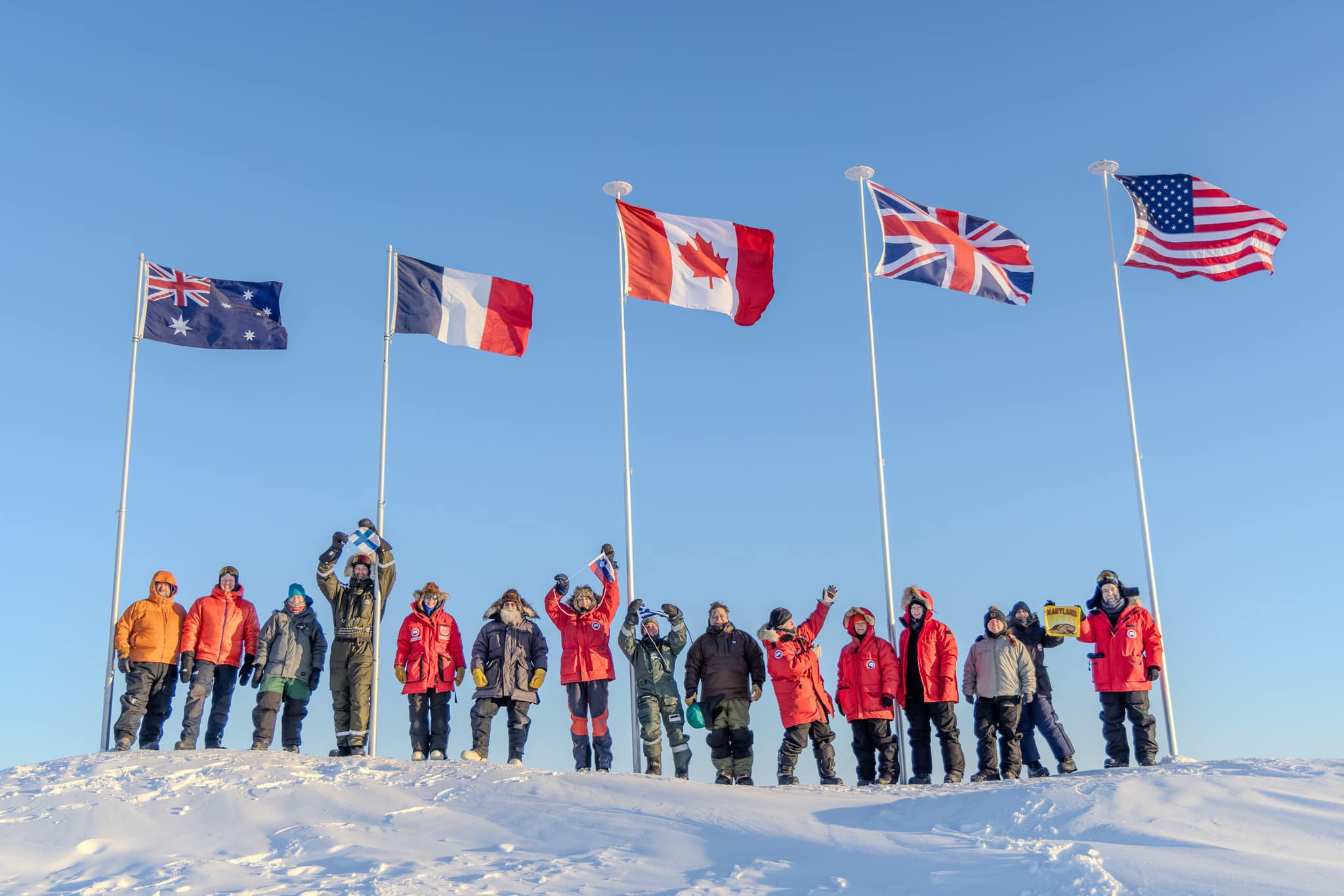 A group of about 15 people bundled in cold-weather gear stand with flags for their countries Australia, France, Canada, United Kingdom, and USA that were planted in Arctic sea ice.