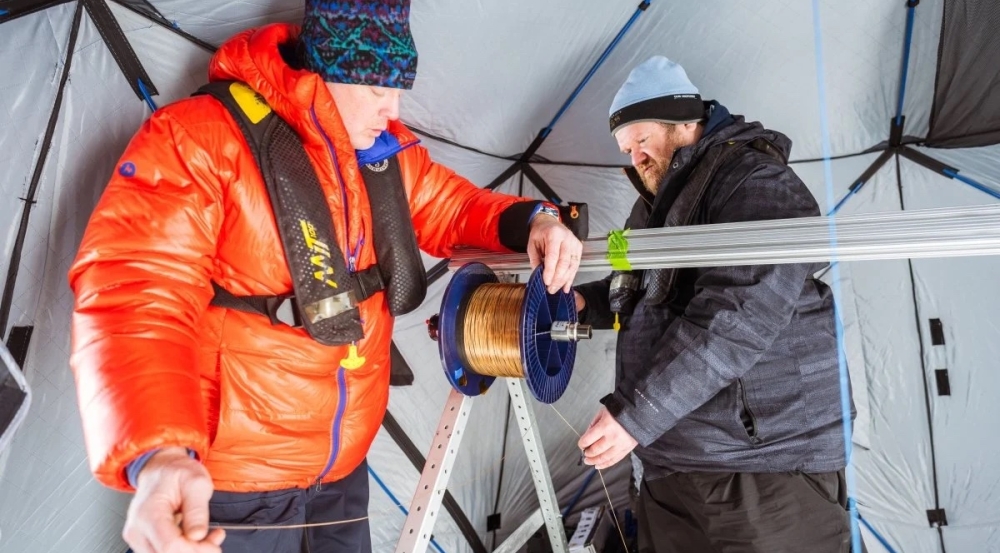 Two people wearing cold-weather gear inspect a fiber spool inside a tent. 