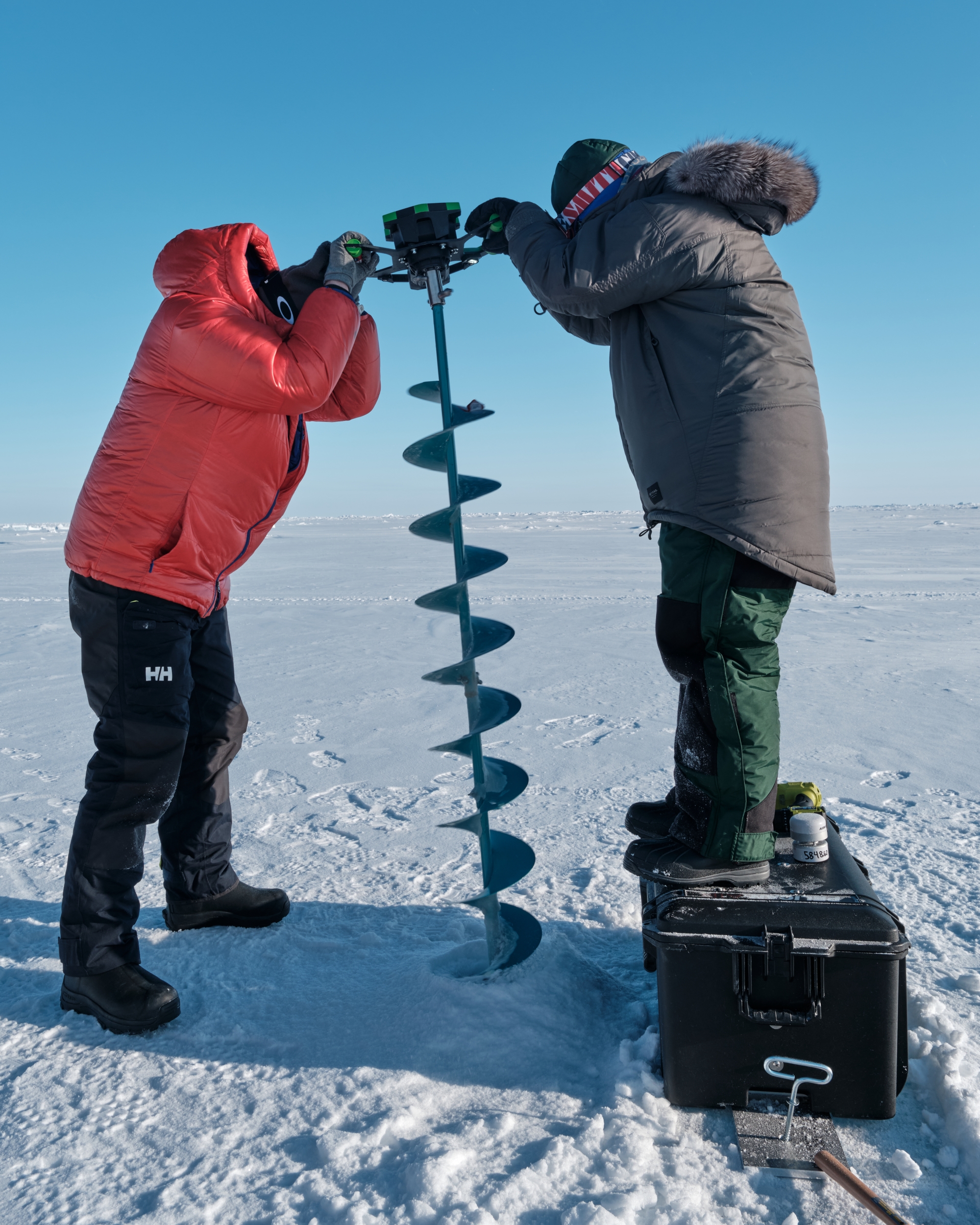 Two people use an auger to drill a hole into sea ice.