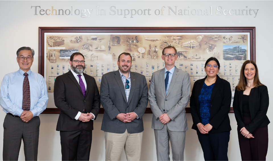 A group of six individuals stand together in front of a plaque displaying technologies developed in support of national security.