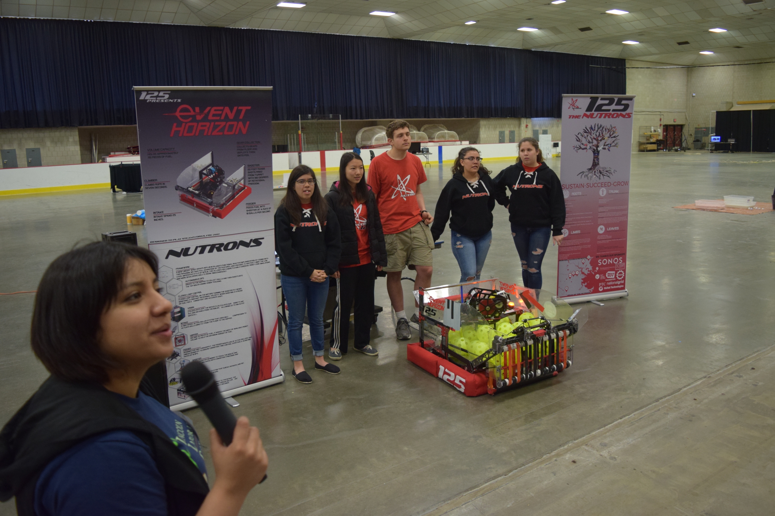 A five-member team stands with a robot as a woman speaks into a microphone.