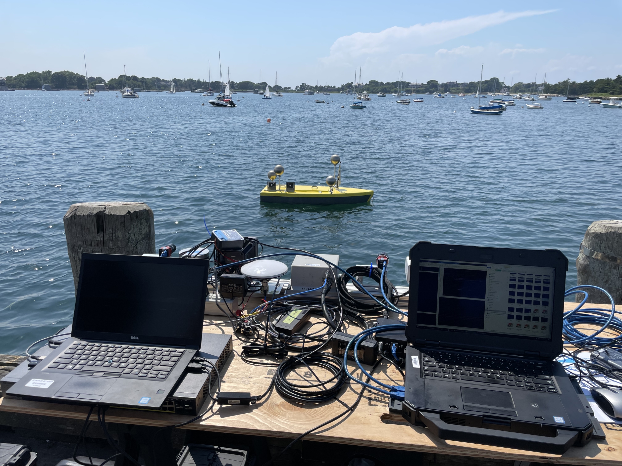 Two laptops are set up on a dock overlooking a harbor with an autonomous surface vessel and sailboats in the background.