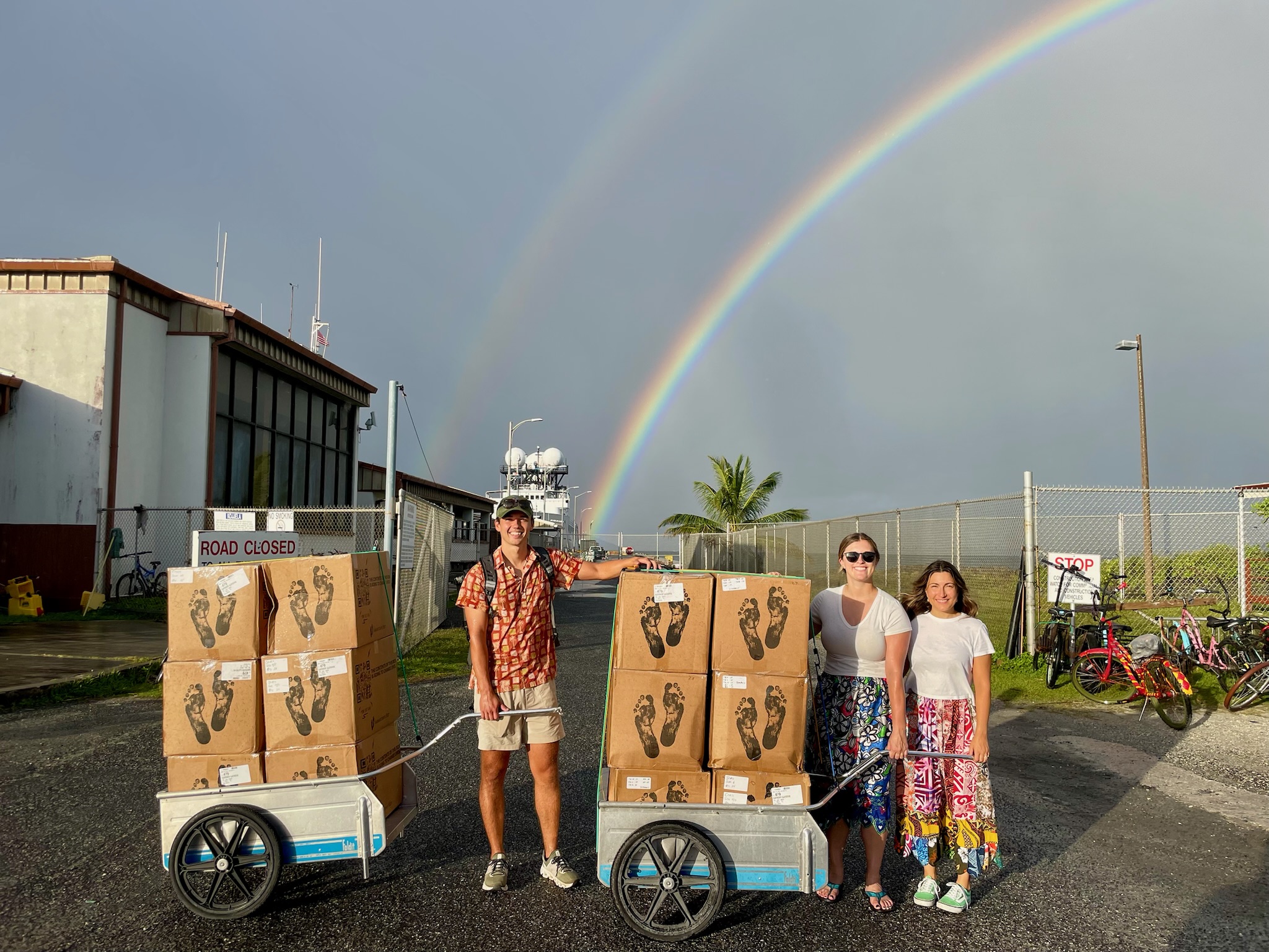 Three people stand outdoors next to carts filled with boxes of donated shoes. A rainbow appears in the background sky.