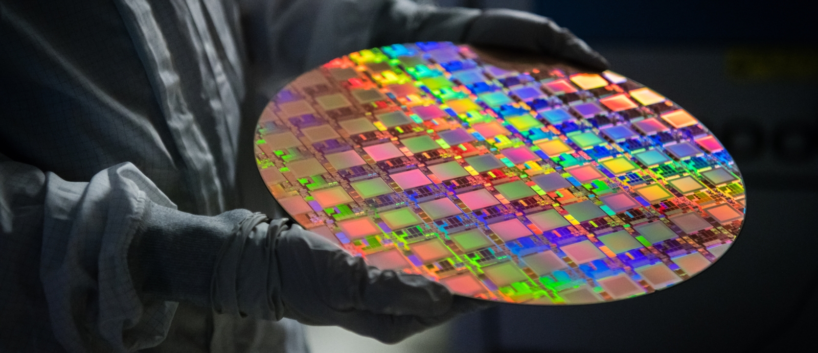 A person with a protective gown and gloves holds a large wafer of integrated circuits. the wafer is vividly multicolored.