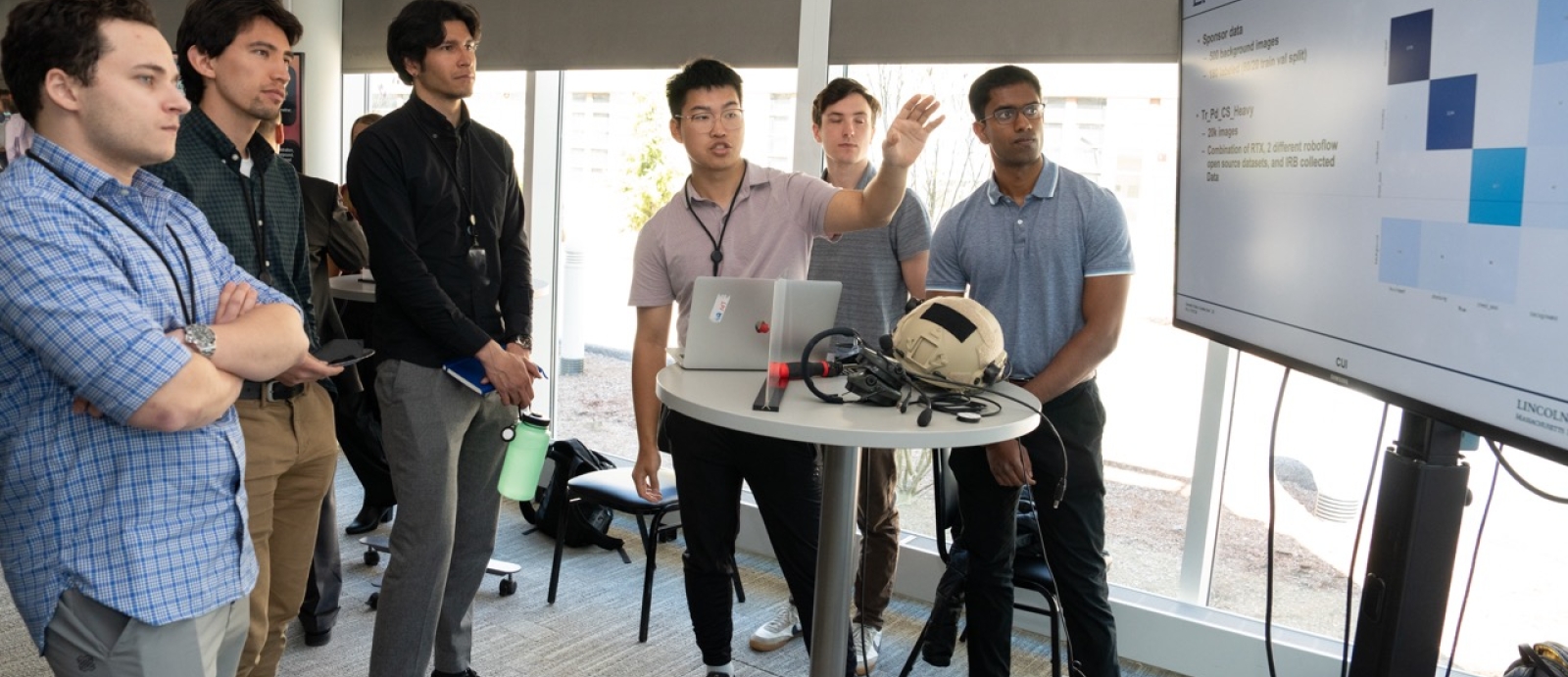 A group of cadets and staff look at a large monitor as a cadet presents his AI casualty status tool.