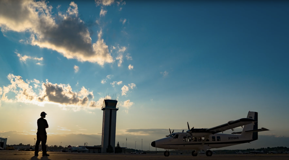 A person standing on a runway with a small aircraft and an air traffic control tower in the background. 