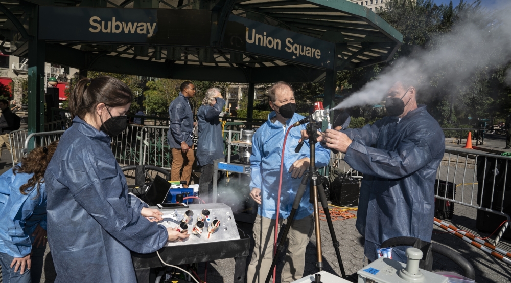 Six scientists in blue lab coats stand in NYC, under a metal gazebo that says "Subway, Union Square" Two of the scientsits operate a small sprayer, on a tripod, that is shooting out a plume of white particles.