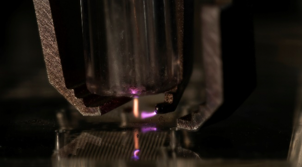 A zoomed-in view of a metal nozzle shooting a stream of orange onto a metal surface, and a nozzle shooting a stream of purple at the orange stream of material. 