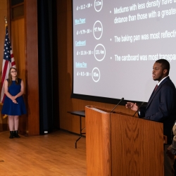 Four high school students stand on an auditorium stage giving a presentation. 