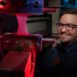 Steve Gillmer is shown smiling next to the industrial knitting machine in red and blue light. 