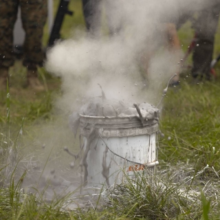 A bucket with silver liquid and gas frothing out of it, on a grassy field.